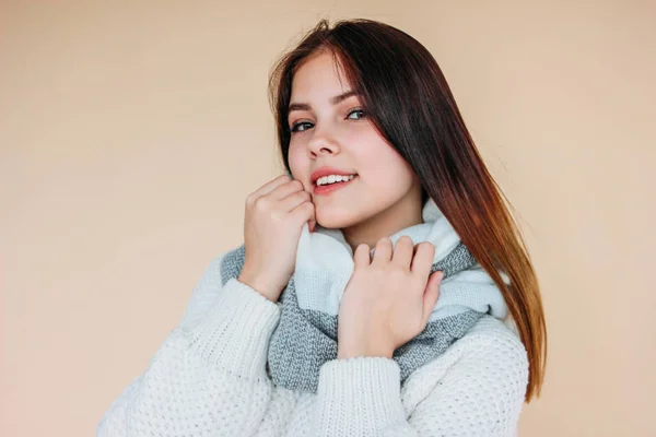 Menina sorridente bonita com pele limpa e cabelos longos escuros em suéter branco acolhedor e cachecol quente no fundo bege — Fotografia de Stock