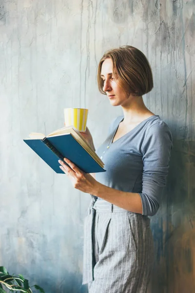 Junge Frau mit gelbem Becher steht an Wand und liest Buch im Kunstatelier, vertikales Porträt — Stockfoto