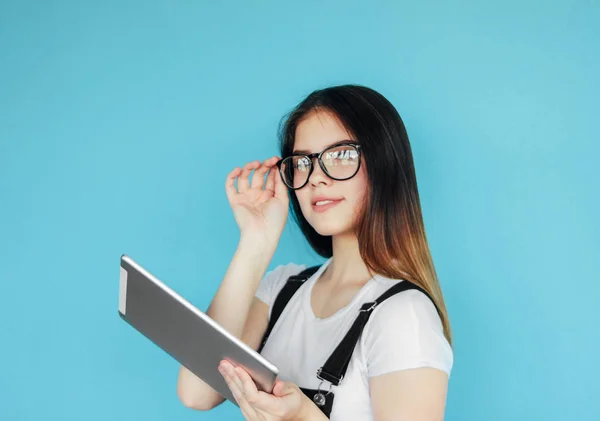 Smailovat krásnou brunetu dívčí teenagera v brýlích a bílých trička držící tabletu v ruce, izolovaná na modrém pozadí — Stock fotografie