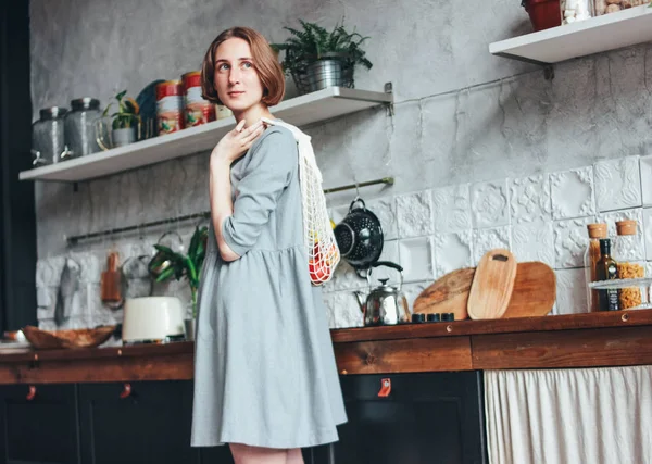 Mujer joven en vestido gris con bolsa de trapo de punto bolsa de hilo comprador en la cocina, cero residuos, la vida lenta — Foto de Stock