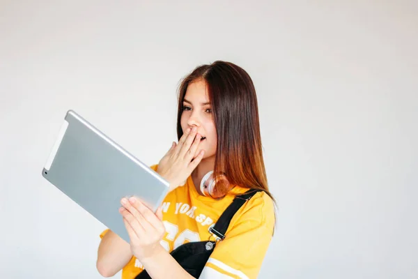 Krásná překvapena brunetka, teenagera ve žlutém tričku, držící tabletu v ruce, izolovaná na bílém pozadí — Stock fotografie