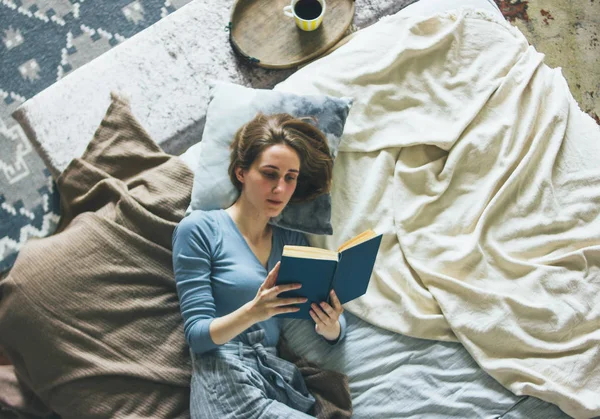 Junge Frau liest Buch auf dem Bett im Dachgeschoss, langsames Leben — Stockfoto
