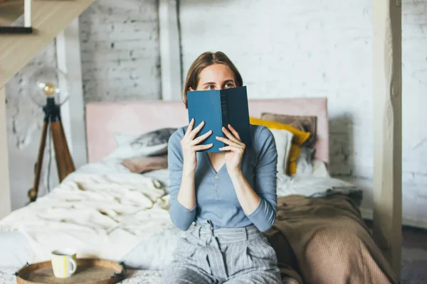 Junge Frau in der Nähe Gesicht Buch sitzt auf dem Bett im Loft Kunstwerk Atelierraum — Stockfoto