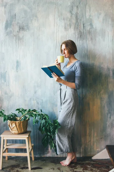 Молода жінка з жовтою чашкою стоїть на стіні і читає книгу в художній майстерні, вертикальний портрет — стокове фото