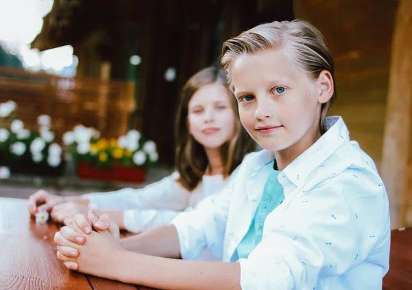 Attractive preteen blonde boy with blue eyes in white shirt with sister friend sitting at table in backyard of wooden cottage — Stock Photo, Image