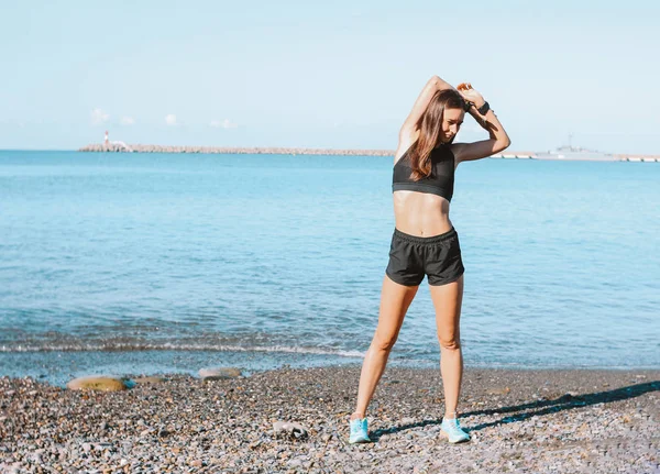 Молодая стройная спортсменка в спортивной одежде делает упражнения на морском пляже утром, здоровый образ жизни — стоковое фото