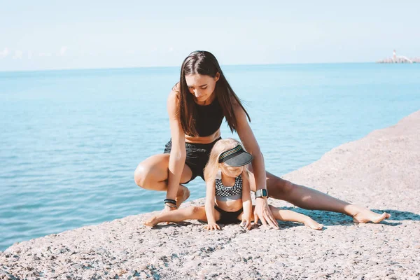 Young fit woman mom with little cute girl exercising on morning beach together, healthy lifestyle, sport family