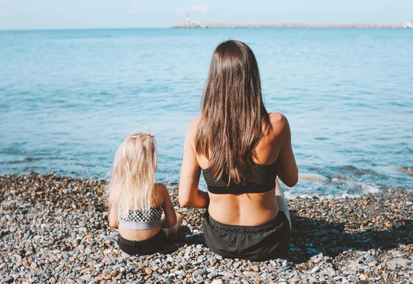 Young fit woman mom with little cute girl sitting on beach together, healthy lifestyle, sport family