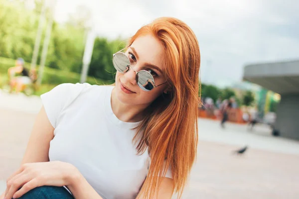 Aantrekkelijke Redhead glimlachend meisje in ronde zonnebril in Casual kleding zittend op straat in de stad — Stockfoto