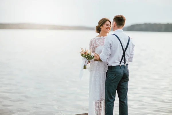 Feliz casal recém-casado perto do lago, sorrindo noiva morena jovem com buquê de boho, ao ar livre — Fotografia de Stock