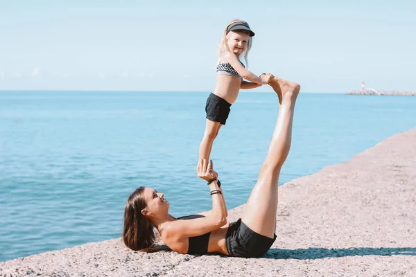 Young fit woman mom with little cute girl exercising on the morning  beach together, healthy lifestyle, sport family