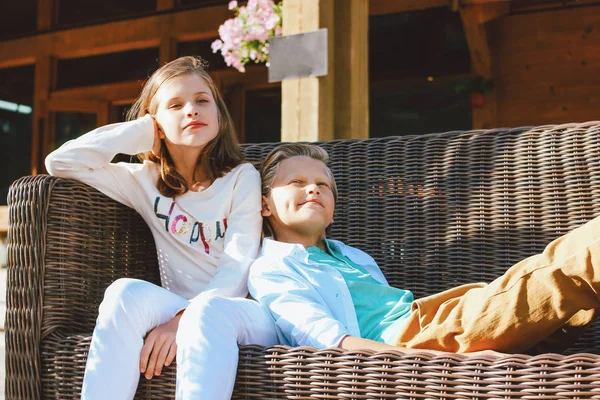 Attractive preteen children sister and brother, friends sitting on wicker sofa in backyard of wooden cottage — Stock Photo, Image