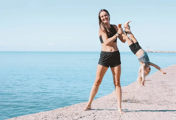 Young fit woman mom with little cute girl exercising on the beach together, healthy lifestyle, dynamic gymnastic