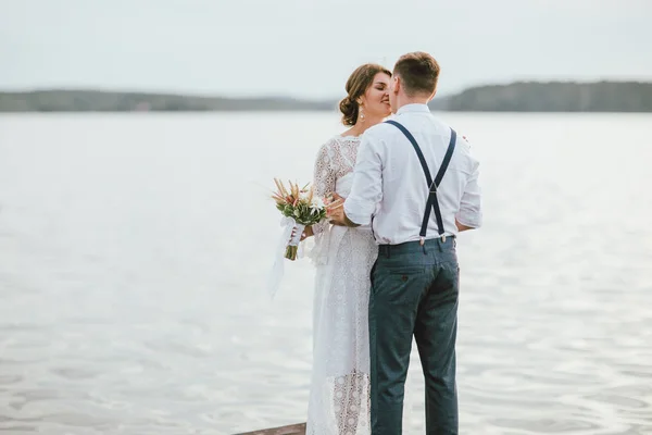 Feliz casal recém-casado perto do lago, noiva morena jovem com buquê de boho, ao ar livre — Fotografia de Stock