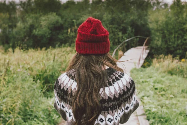 Beautiful carefree long hair girl in the red hat and knitted nordic sweater in autumn nature park, people from behind. Travel adventure lifestyle — Stock Photo, Image