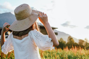 Dağda güzel evlerin arka planına karşı arkadan hasır şapka güzel romantik preteen kız, gün batımında kırsal sahne