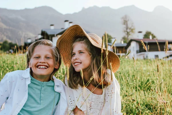 Happy children brother and sister friends sitting in grass against the background of beautiful houses in mountain, family travel, rural scene — Stock Photo, Image