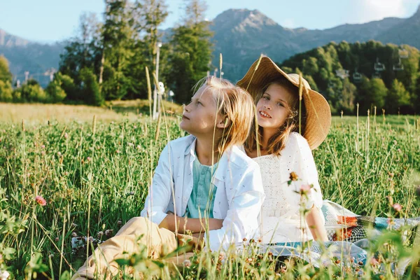 Children brother and sister friends sitting in grass against the background of beautiful houses in mountain, family travel, rural scene — Stock Photo, Image