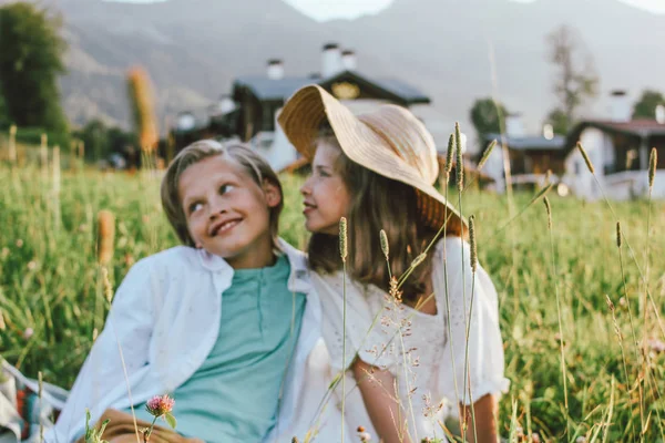 Niños hermanos y hermanas amigos sentados en la hierba contra el fondo de hermosas casas en las montañas. Enfócate en la hierba, modelos borrosos —  Fotos de Stock