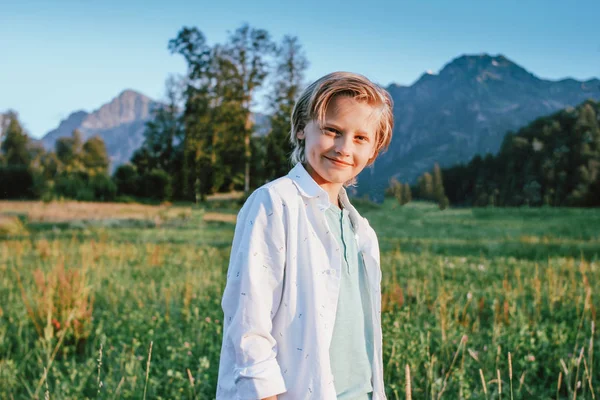Blonďatý usměvavý chlapec, který hledí na kameru na pozadí nádherného pohledu na zelenou louku a hory, rodinné cestování dobrodružný životní styl — Stock fotografie