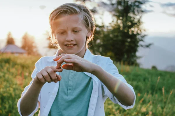 Blonde lachende jongen houdt een sprinkhaan in zijn hand bij zonsondergang weide, landelijke scène — Stockfoto