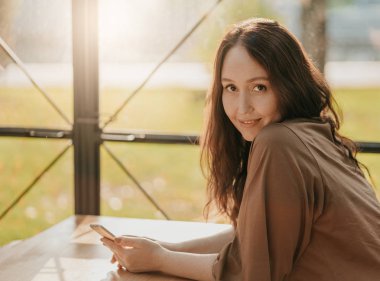 Elinde cep telefonu ile kafede pencerede oturan uzun kıvırcık saçlı Dostu büyüleyici esmer kadın