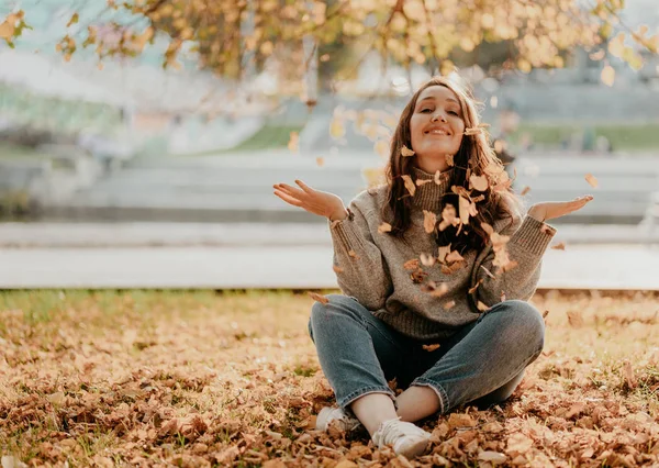 地面に座って長い巻き毛を持つフレンドリーな魅力的なブルネットの女性と黄金の葉を投げる、秋の季節 — ストック写真