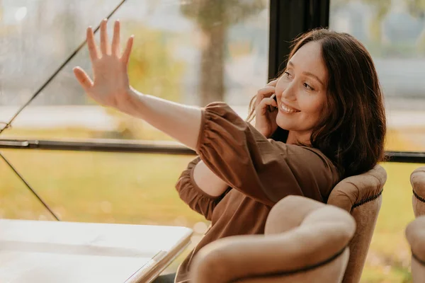 Vriendelijke charmante brunette vrouw met lang krullend haar zittend in het raam in café met mobiele telefoon en zwaaien naar vrienden — Stockfoto