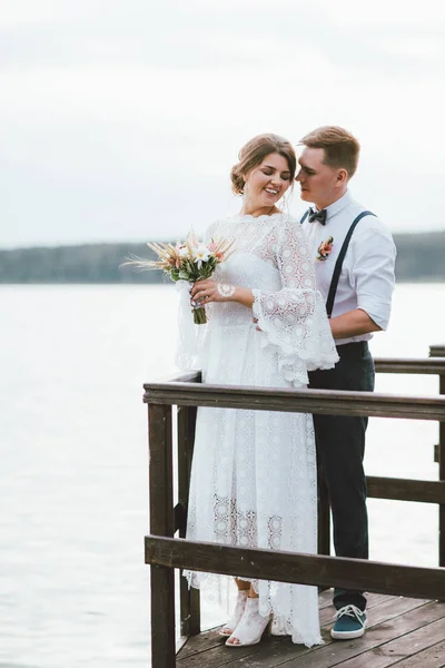 Feliz casal recém-casado, sorrindo noiva morena jovem com buquê estilo boho olhando para o noivo, ao ar livre — Fotografia de Stock