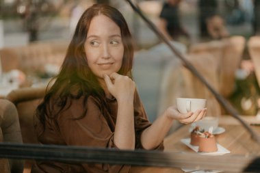 Elinde kahve fincanı ile kafede pencerede oturan uzun kıvırcık saçlı Büyüleyici esmer kadın