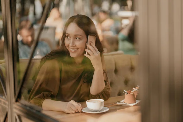 Charming brunette woman with long curly hair sitting at the window in cafe with mobile phone in hands