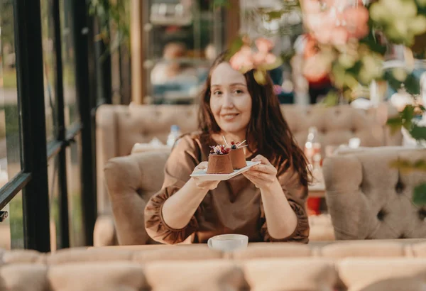 Очаровательная брюнетка с длинными вьющимися волосами сидит у окна в кафе с десертом в руках, избирательный фокус — стоковое фото