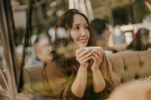Очаровательная брюнетка с длинными вьющимися волосами сидит у окна в кафе с чашкой кофе в руках — стоковое фото