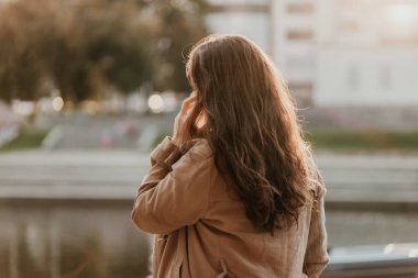 Uzun kıvırcık saçlı arkadan büyüleyici gülümseyen esmer kadın şehir caddesinde elinde cep telefonu ile rahat ceket giymiş