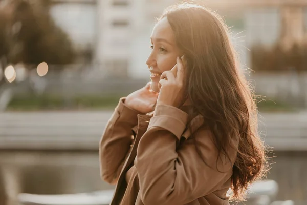 Γοητευτική χαμογελαστή μελαχρινή γυναίκα με μακριά σγουρά μαλλιά ντυμένη περιστασιακή παλτό με το κινητό τηλέφωνο στα χέρια στο δρόμο της πόλης — Φωτογραφία Αρχείου