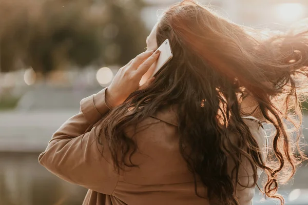 Mulher morena sorridente encantadora com cabelo encaracolado longo ventoso vestido casaco casual com telefone celular nas mãos na rua da cidade — Fotografia de Stock