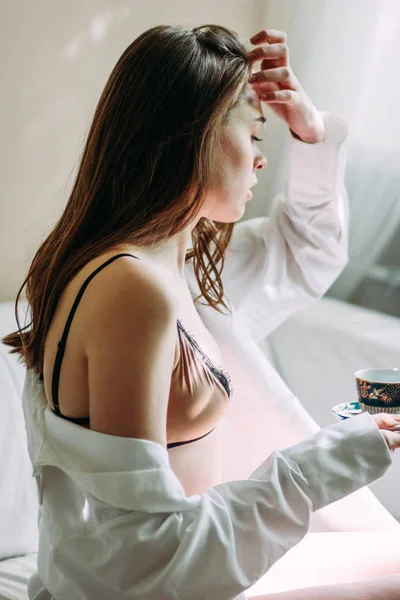 Sensual muda cantik gadis rambut panjang model pakaian dalam dan kemeja putih duduk di tempat tidur dengan secangkir teh pagi, lembut fokus — Stok Foto