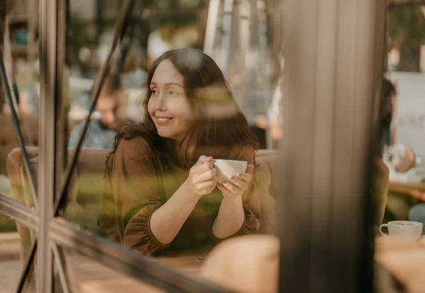 Очаровательная брюнетка с длинными вьющимися волосами сидит у окна в кафе с чашкой кофе в руках — стоковое фото