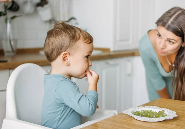 Charming concentrated little baby boy eating first food green grape with mother at the bright kitchen at home