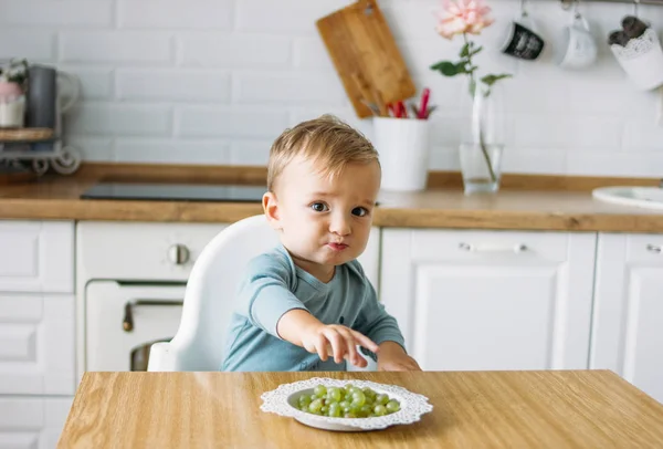 Очаровательный маленький мальчик ест первую еду зеленый виноград на яркой кухне дома — стоковое фото