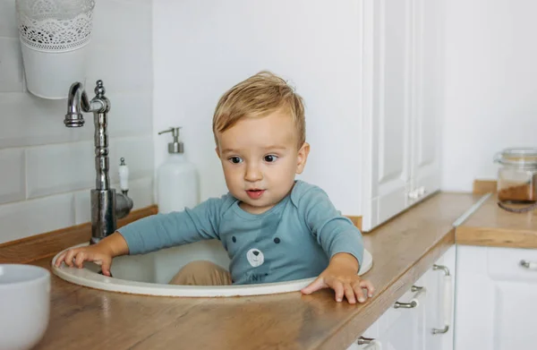 Очаровательный маленький мальчик сидит в раковине на яркой кухне дома — стоковое фото