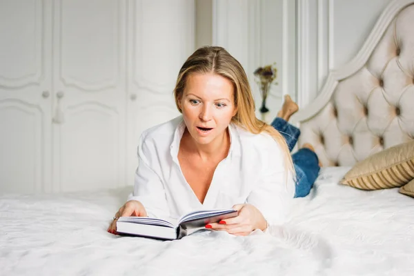 Mulher loira encantadora surpresa com cabelo longo e claro em roupas casuais livro de leitura na cama em interior rico brilhante — Fotografia de Stock