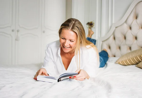 Mulher loira encantadora feliz com cabelo longo e claro em roupas casuais livro de leitura na cama em interior rico brilhante — Fotografia de Stock
