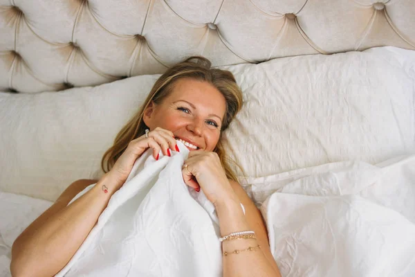 Happy tersenyum indah panjang wanita rambut adil mengenakan pakaian dalam menutup wajah duduk di tempat tidur di interior yang kaya cerah — Stok Foto