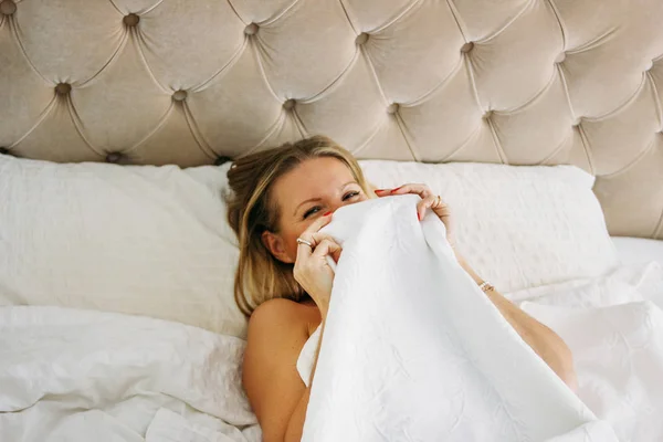 Happy tersenyum indah panjang wanita rambut adil mengenakan pakaian dalam menutup wajah duduk di tempat tidur di interior yang kaya cerah — Stok Foto