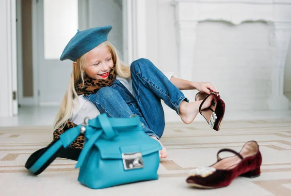Cute fashion trendy fair hair little girl dressed in denim overalls puts on shoes, leopard scarf and blue beret with mom's accessories. Daughter imitates mom — Stock Photo, Image