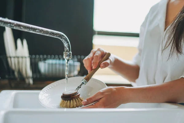 Young woman washes dishes with wooden brush with natural bristles at window in kitchen. Zero waste concept