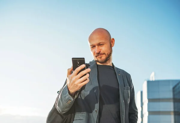 Handsome adult bald bearded man businessman in suit using mobile phone on background of blue sky and glass modern building