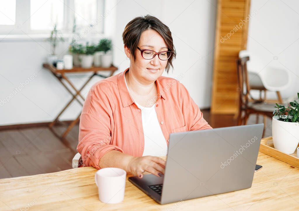 Adult charming brunette woman in glasses plus size body positive working with laptop at home office