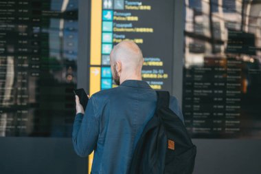 Sırt çantalı, kel, takım elbiseli adam havaalanında bilgi okuyor. 